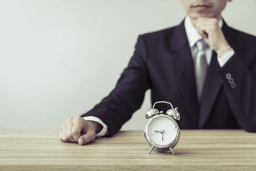 中国 スーツを着た男性が目覚まし時計を持ってテーブルに座っている。 中国 スーツを着た男性が目覚まし時計を持ってテーブルに座っている。