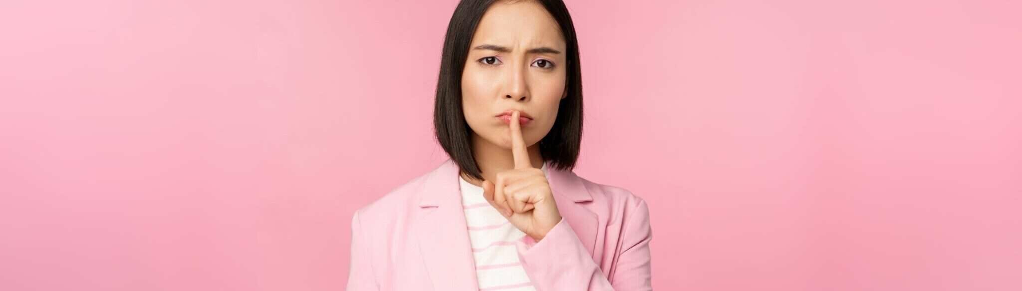 ピンクのスーツを着た女性が唇に指を当て、沈黙と秘密を象徴している。