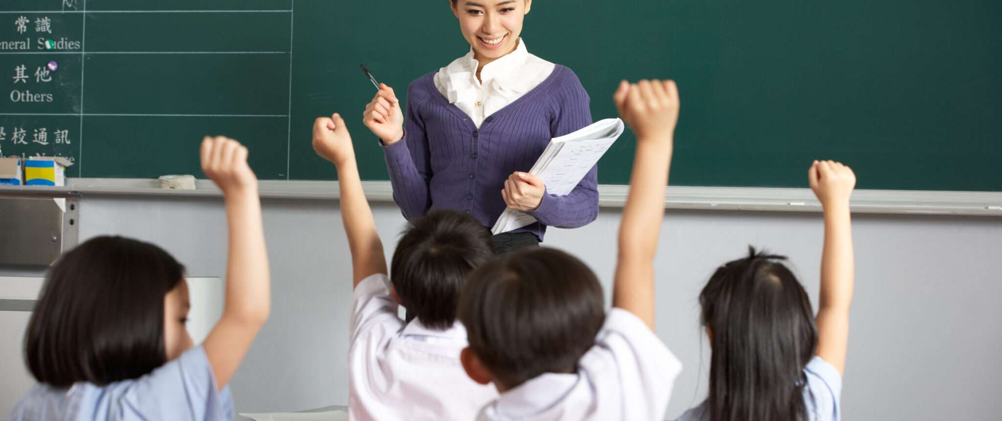 中国の教師が教室の前に立っていて、子供たちが手を挙げています。