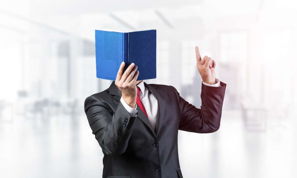 中国の明るいオフィス空間で、青い本で顔を覆い、上を向いているスーツを着たビジネスマン。