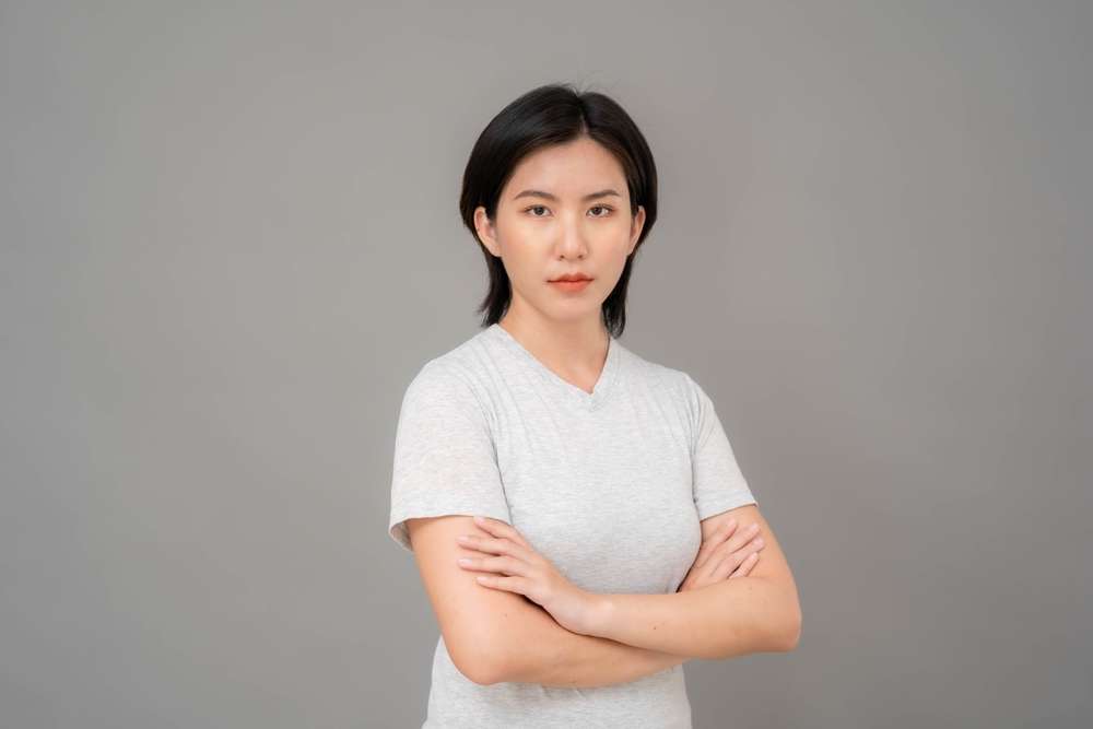 中国で、白いTシャツを着た黒髪の女性が腕を組んで立ち、灰色の背景を背にカメラをまっすぐ見つめている。