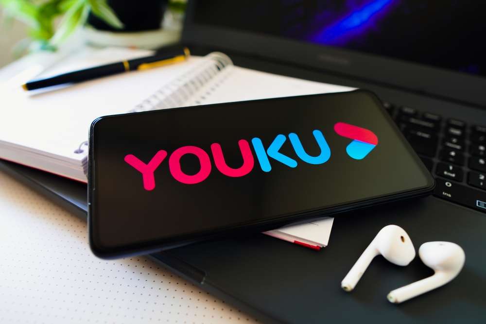 Youku のロゴが表示されたスマートフォンが、白いイヤホン、ノート、ペンの横にあるノートパソコンのキーボードの上に置かれています。これは、中国のトップ ストリーミング コンテンツを楽しむための理想的なセットアップです。 Youku のロゴが表示されたスマートフォンが、白いイヤホン、ノート、ペンの横にあるノートパソコンのキーボードの上に置かれています。これは、中国のトップ ストリーミング コンテンツを楽しむための理想的なセットアップです。