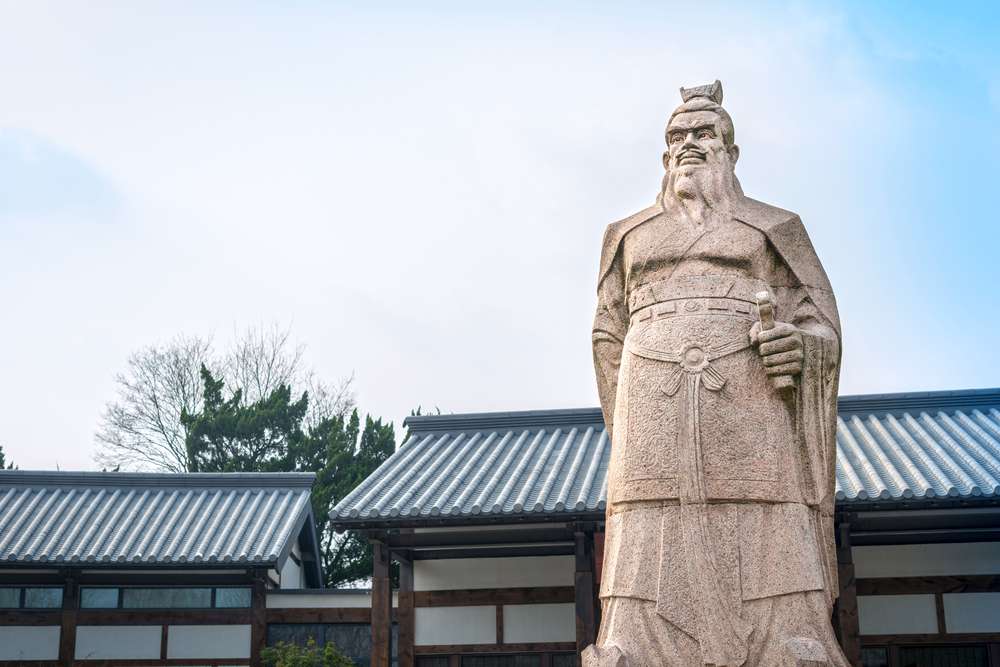 晴れた日に瓦屋根の建物の前に立つ、伝統的な衣装を着た髭を生やした男性の石像は、中国の豊かな文化遺産を思い起こさせます。