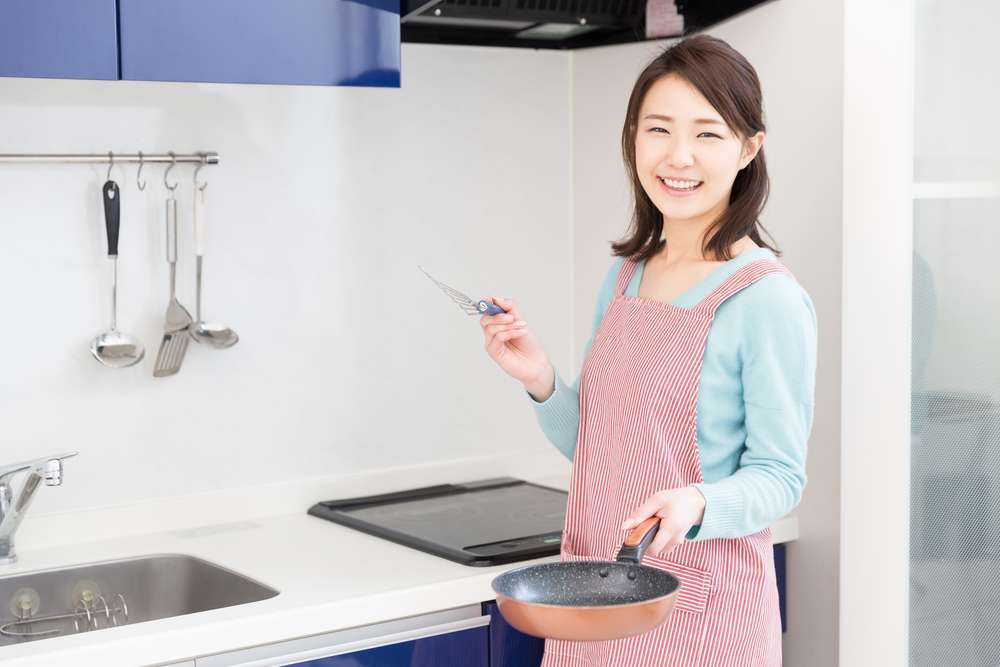 ピンクのエプロンを着た女性がキッチンに立って、フライパンとヘラを持ち、カメラに向かって微笑んでいる。彼女の後ろの壁にはキッチン用品が掛けられており、伝統的な中国のキッチンを彷彿とさせる。 ピンクのエプロンを着た女性がキッチンに立って、フライパンとヘラを持ち、カメラに向かって微笑んでいる。彼女の後ろの壁にはキッチン用品が掛けられており、伝統的な中国のキッチンを彷彿とさせる。