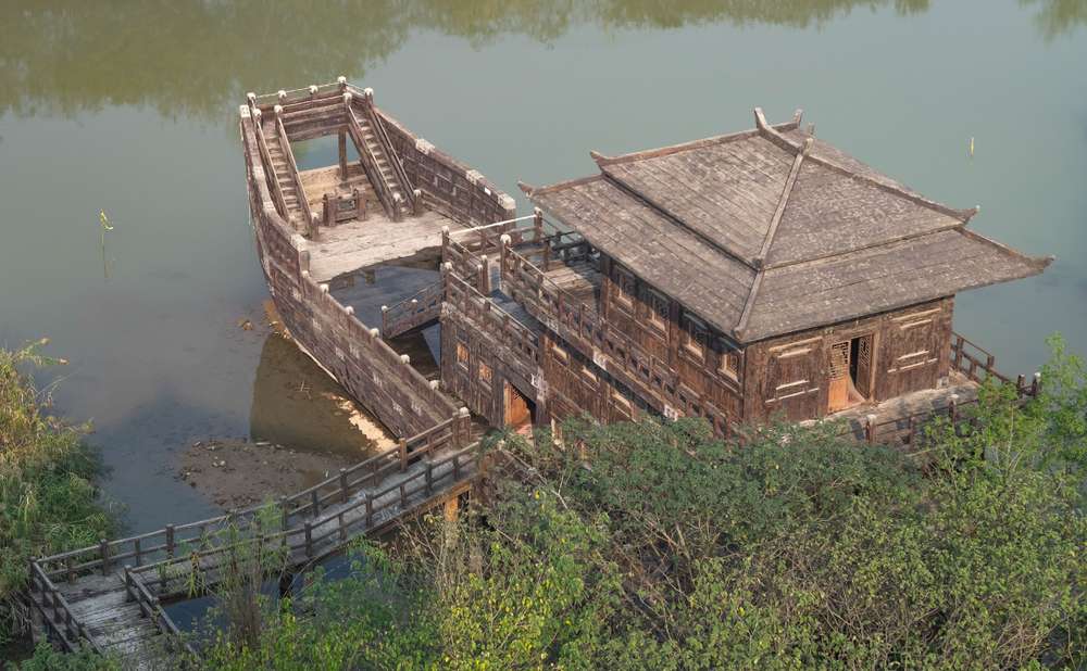 伝統的な船のようなデザインの木造建築物が、緑に囲まれた静かな水辺に停泊しており、中国の穏やかな風景を彷彿とさせる雰囲気を醸し出しています。 伝統的な船のようなデザインの木造建築物が、緑に囲まれた静かな水辺に停泊しており、中国の穏やかな風景を彷彿とさせる雰囲気を醸し出しています。