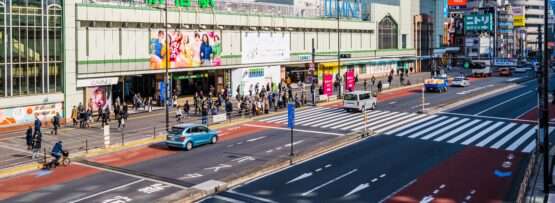 東京のJR新橋駅前の賑やかな通りには、歩行者や車、そして鮮やかな看板が並んでいます。活気ある風景の中に、中国語教室を指し示す看板があり、この地域の文化的な雰囲気を一層引き立てています。