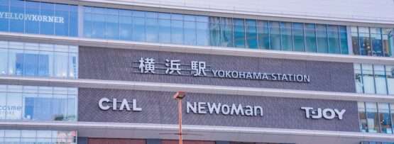 横浜駅の外観は、CIAL、NEWoMan、TJOYの看板が並び、大きな窓とレンガ造りのファサードが横浜の活気あるエネルギーを表現しています。