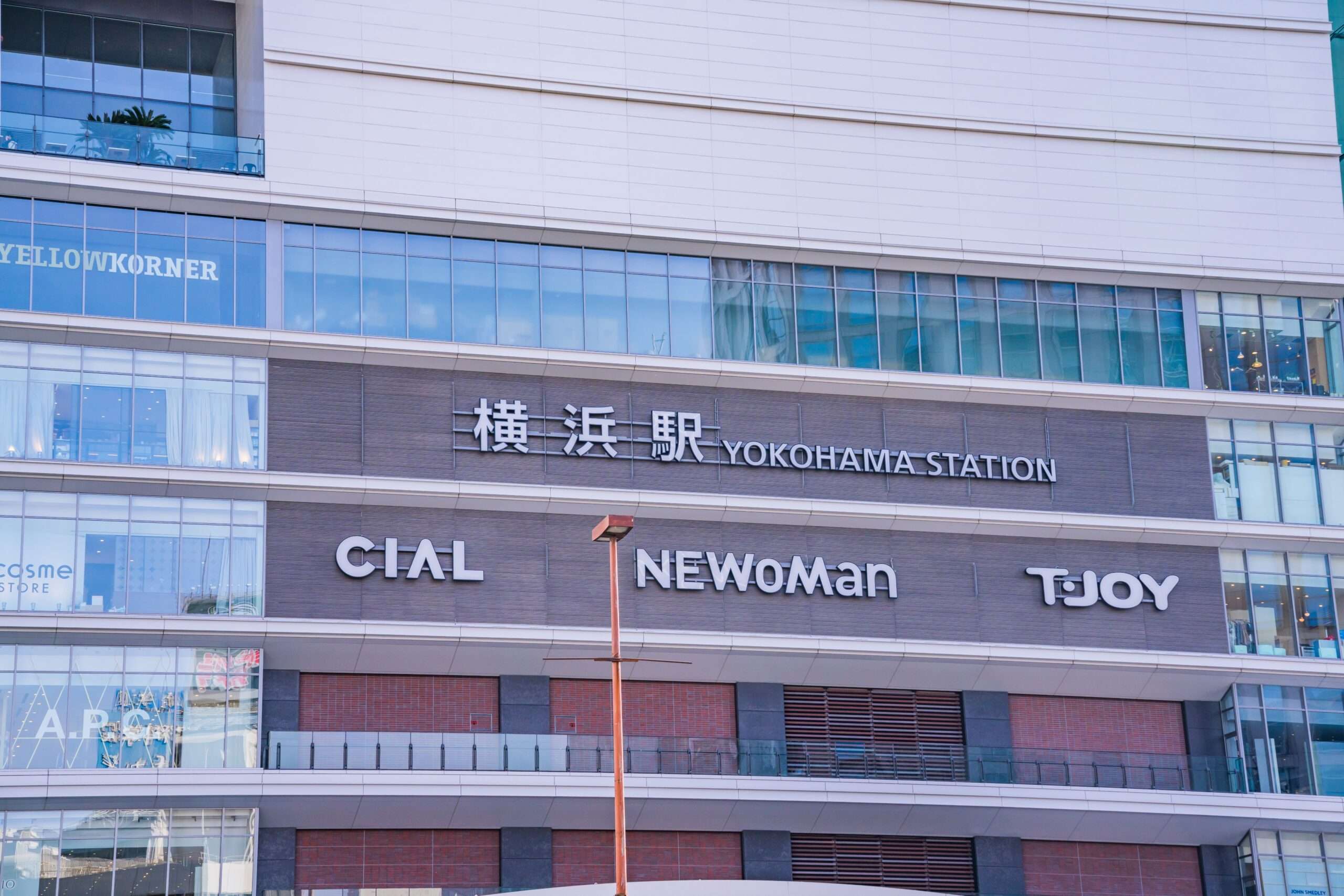 横浜駅の外観は、CIAL、NEWoMan、TJOYの看板が並び、大きな窓とレンガ造りのファサードが横浜の活気あるエネルギーを表現しています。