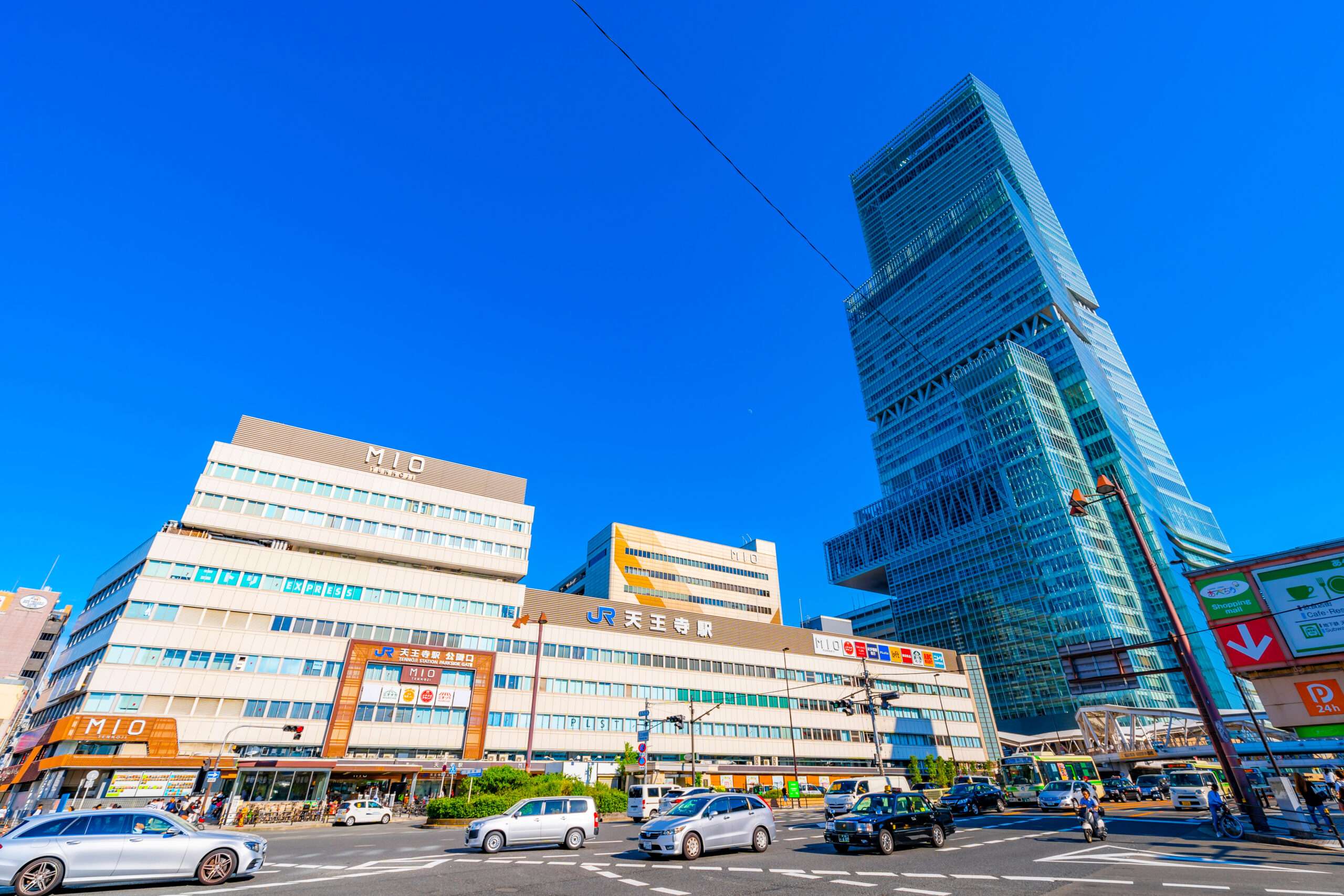 天王寺近くの賑やかなショッピング センターと混雑した交差点の隣にある近代的な高層ビルの街並み。澄んだ青空の下、車や歩行者が活発に動いています。