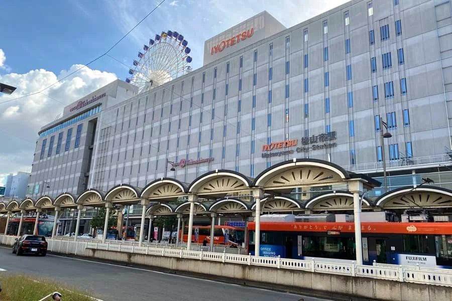 一部曇り空の下、「NORETSU」の看板が掲げられた大きなビル、正面にはタクシー乗り場、背景には観覧車が見える。.