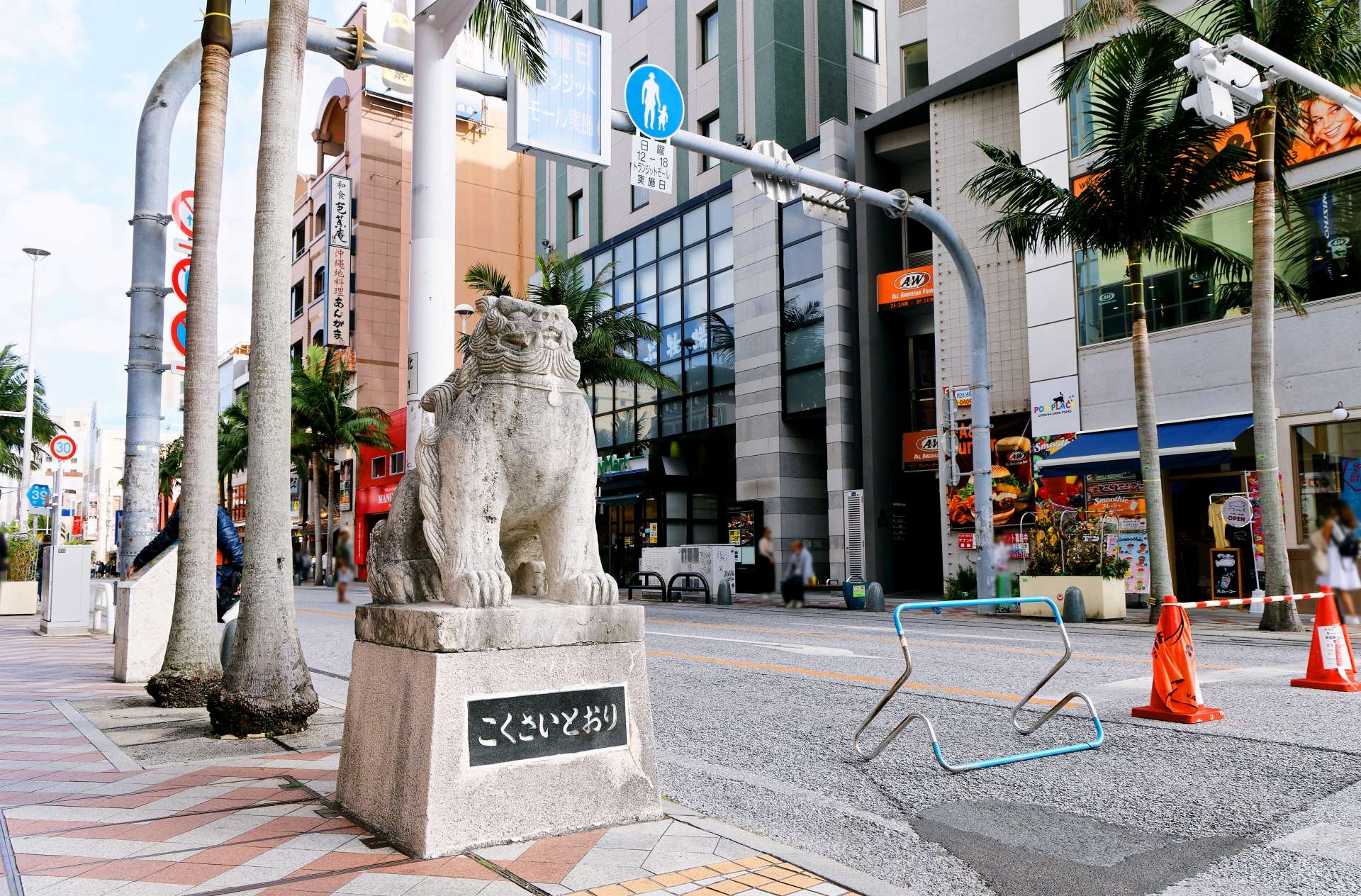 沖縄の那覇市の賑やかな街角に石造りのライオン像が立っており、建物やヤシの木、交通標識に囲まれている。近くには中国語教室を指し示す標識もある。