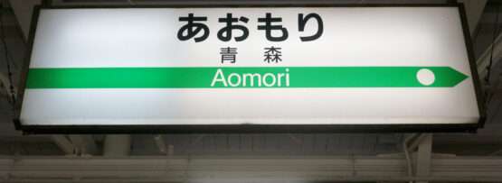 緑の路線線と方向矢印とともに日本語と英語で「Aomori」と書かれた駅の標識が、青森・弘前へ旅する旅行者を歓迎します。