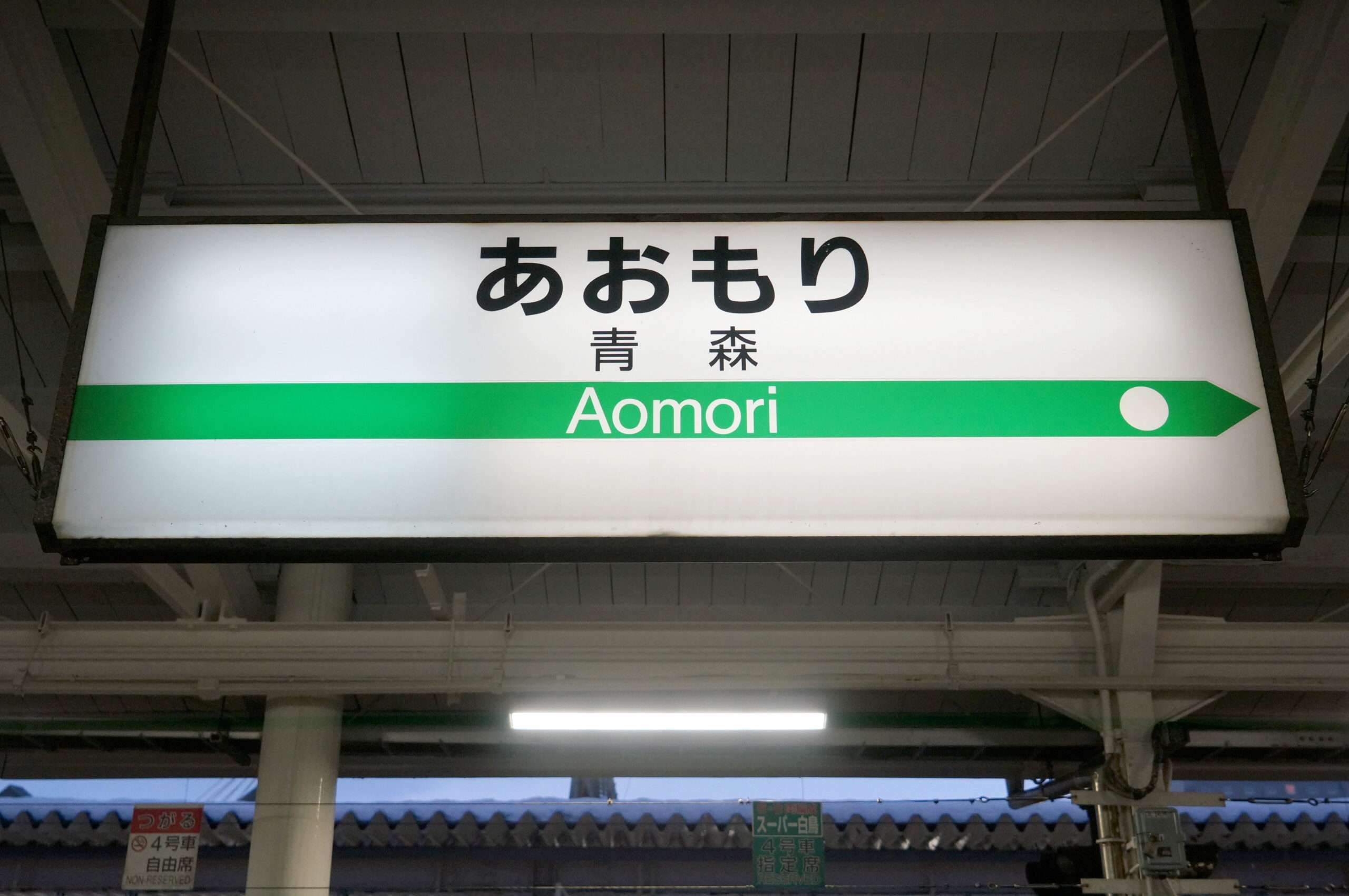 緑の路線線と方向矢印とともに日本語と英語で「Aomori」と書かれた駅の標識が、青森・弘前へ旅する旅行者を歓迎します。