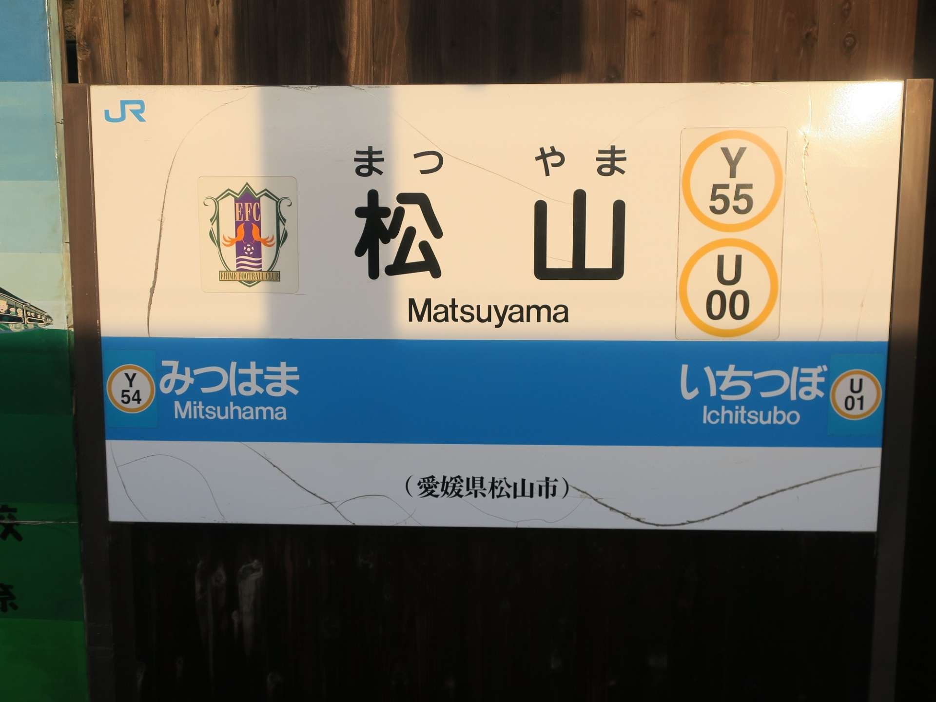 松山 (愛媛・松山) の鉄道駅標識。Y55 と U00 の路線マーカーと、追加の停留所三津浜 (Y54) と市坪 (U01) を示しています。