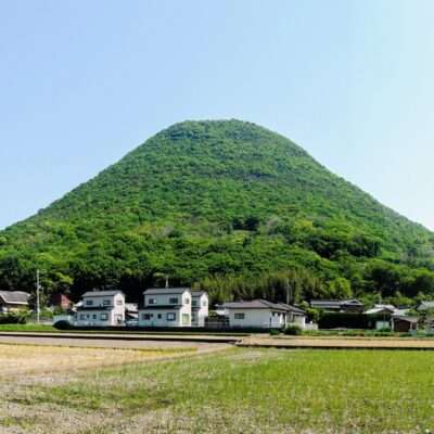 香川・高松の背景には緑豊かな丘がそびえ立ち、手前には澄み切った青空の下、小さな家々と平らな畑が広がっています。近くには活気ある中国語学校があり、この絵のように美しい風景の中で文化交流が活発に行われています。