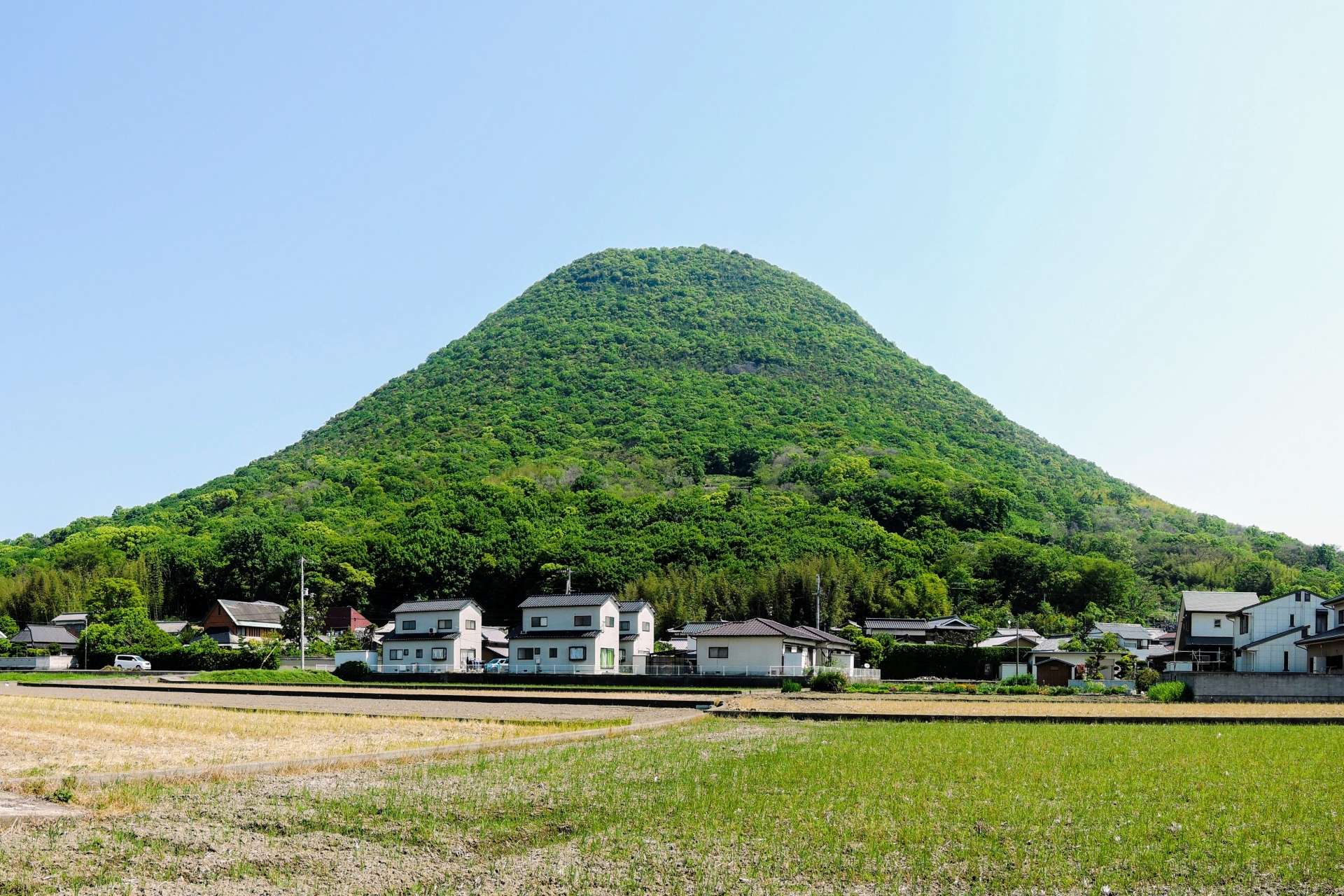 香川・高松の背景には緑豊かな丘がそびえ立ち、手前には澄み切った青空の下、小さな家々と平らな畑が広がっています。近くには活気ある中国語学校があり、この絵のように美しい風景の中で文化交流が活発に行われています。