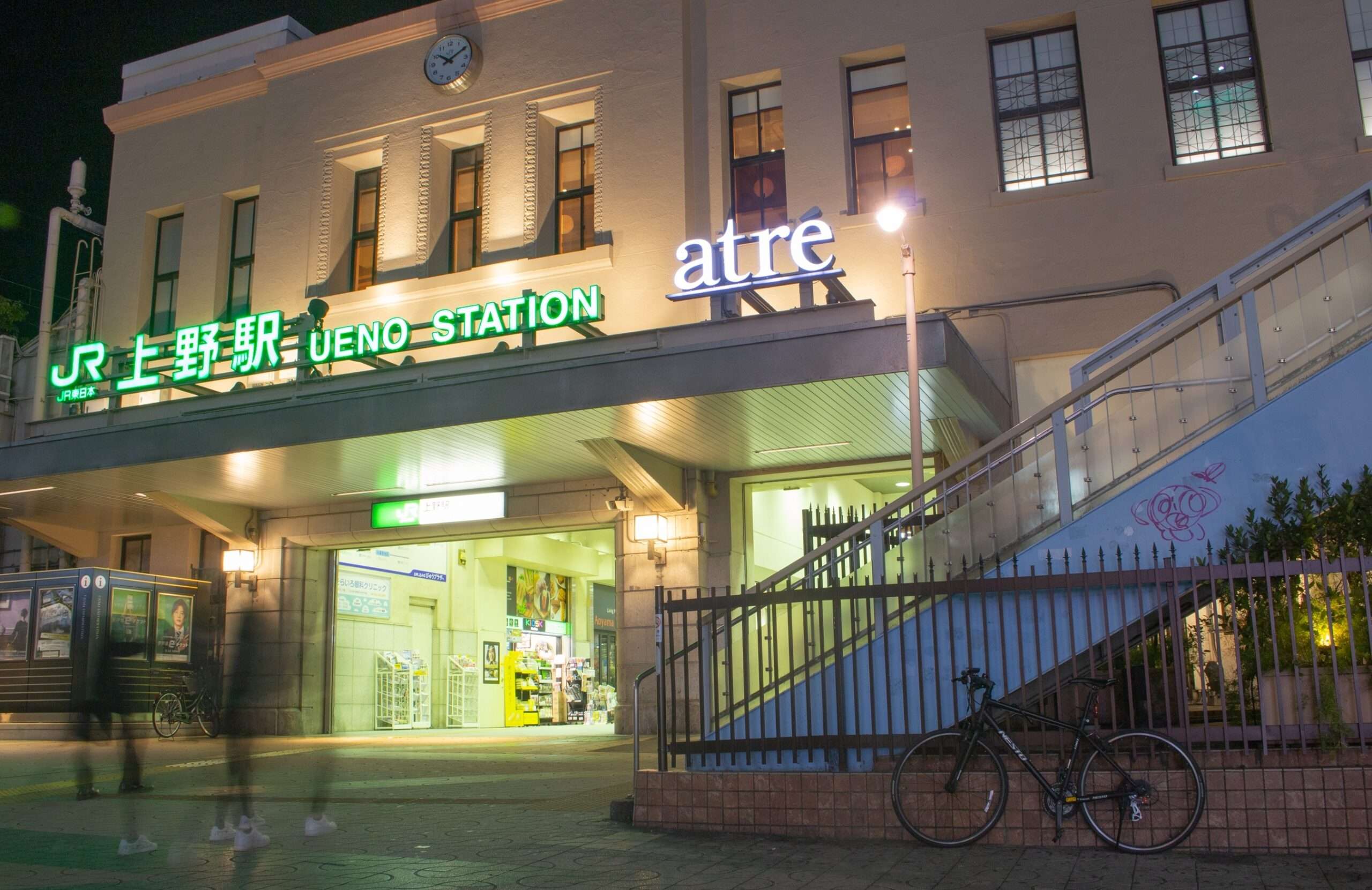 夜になると、緑の看板で飾られた上野駅の賑やかな入り口が静かに佇み、ぼんやりとした人影が通り過ぎていく。近くには自転車が停まっており、時計の付いた階段がその光景に彩りを添えている。近くの中国語教室は、夕方の勉強を続ける熱心な学習者で賑わっている。
