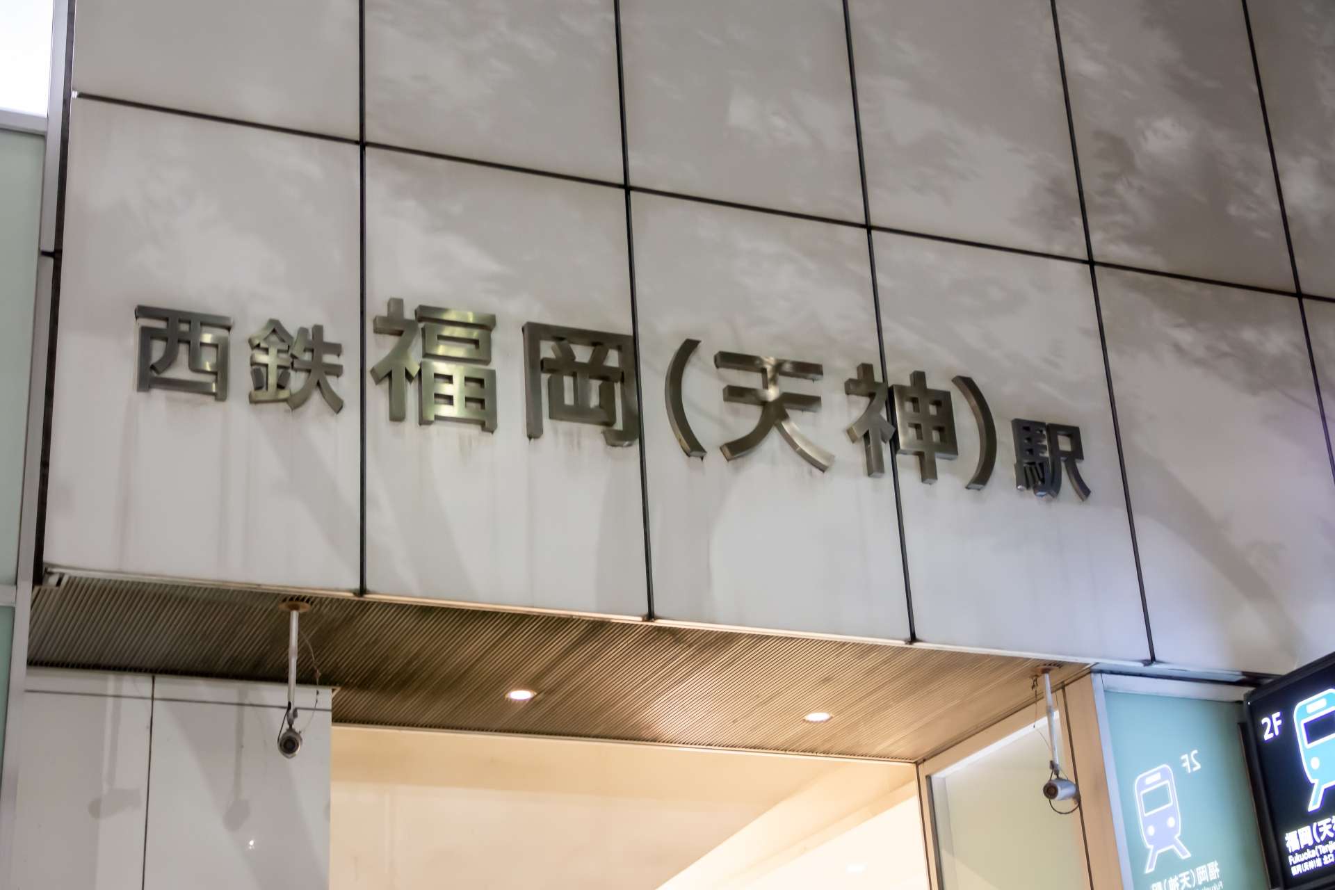 日本の西鉄福岡（天神）駅の入り口にある看板。真っ白なタイル張りの壁に、なめらかな金属の文字が描かれています。近くには、天神の中心で言語と文化が融合する中国語教室があります。