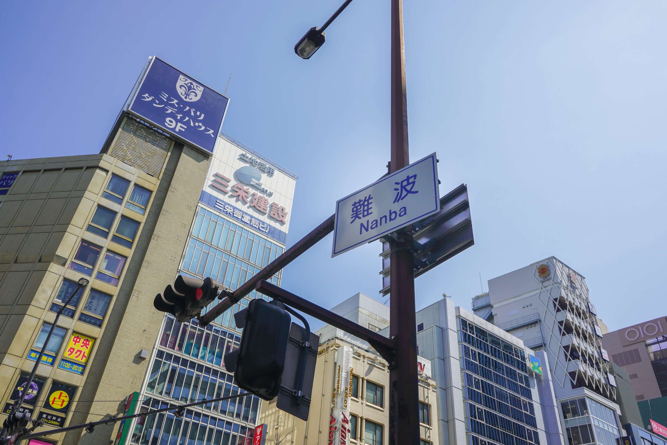 難波のストリート ビュー、難波・なんば。澄んだ青い空の下、信号機と日本の標識で飾られた高層ビルが特徴です。賑やかな通りの中に、中国語を習得するために熱心な学習者が集まる中国語教室があります。