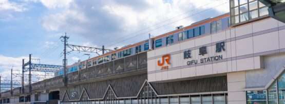 晴れた日に岐阜駅の上の高架線路を列車が走行している。背景の空は部分的に曇っており、近くの建物に影が落ちている。