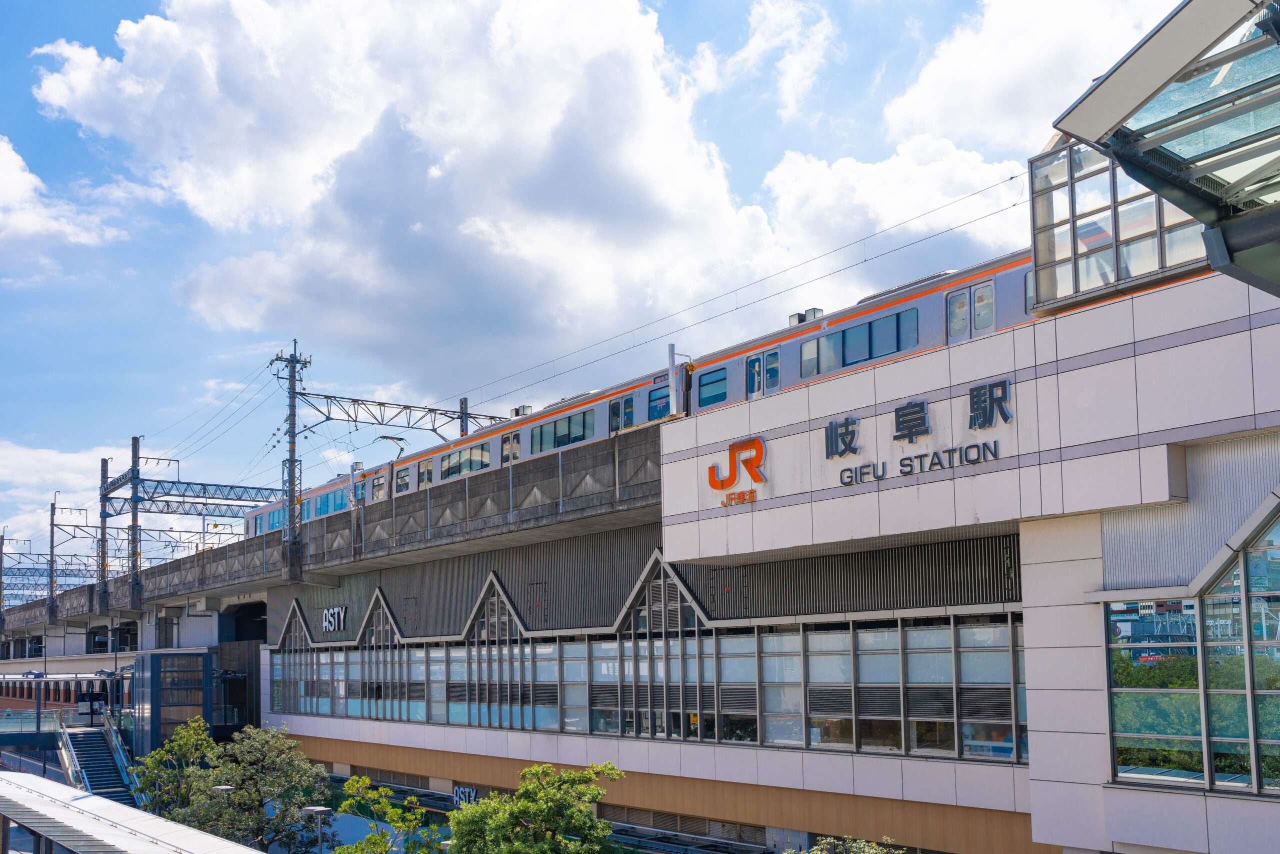 晴れた日に岐阜駅の上の高架線路を列車が走行している。背景の空は部分的に曇っており、近くの建物に影が落ちている。