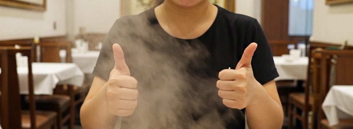 レストランで、湯気がたつ餃子のかごの後ろで、笑顔の人が両手の親指を立てている。.