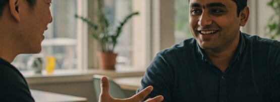 明るいカフェのテーブルに二人の男性が座り、会話に花を咲かせている。一人が手振りをしながら、もう一人は微笑みながら話を聞いている。.