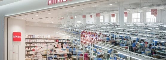 左側に商品が展示されている MINISO の店頭、右側には従業員がいる明るく照らされた大きな工場または倉庫。.
