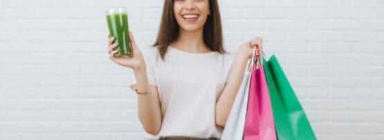 白いレンガの壁を背に立つ女性。片手にカラフルな買い物袋をいくつも持ち、もう片方の手には緑色の飲み物を持ち、カメラに向かって微笑んでいる。.