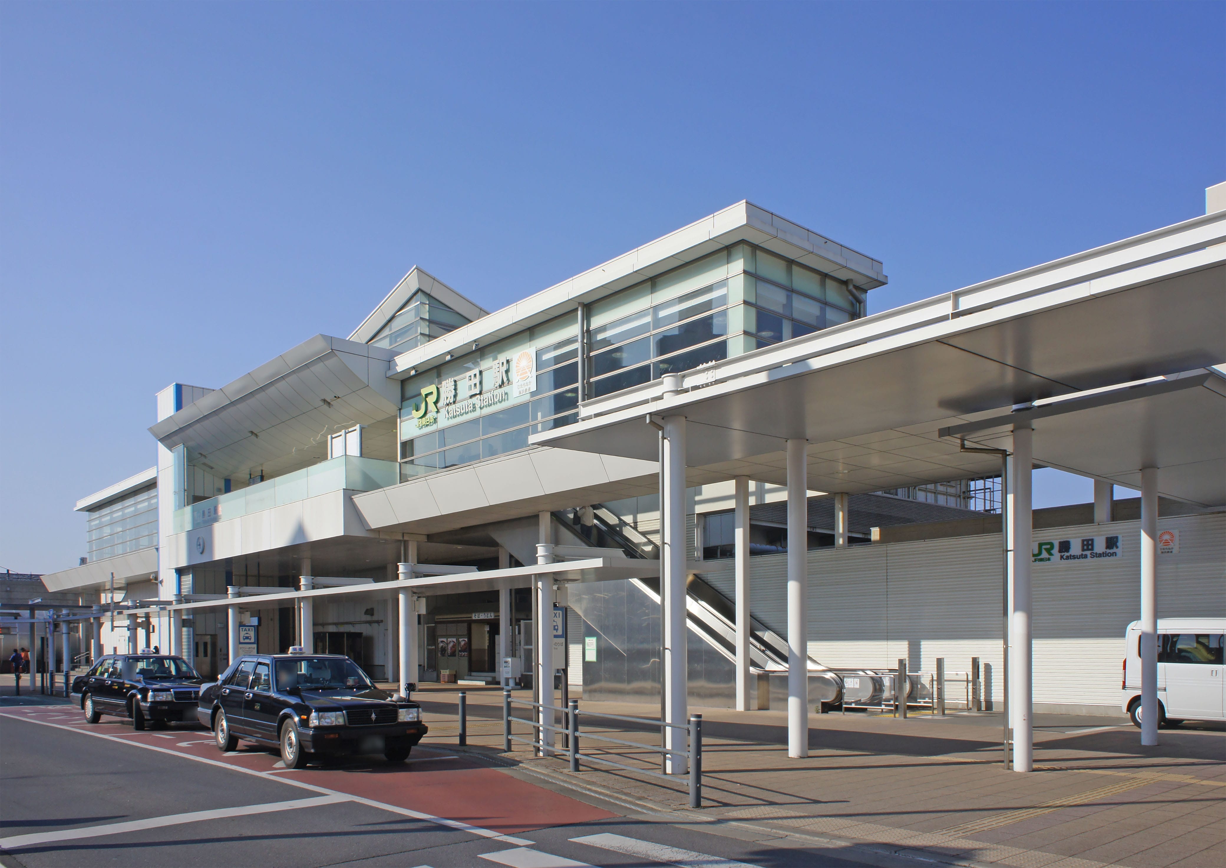 ガラス張りの近代的な駅舎、高架の歩道、正面に停まるタクシー、頭上には晴れ渡った空。.