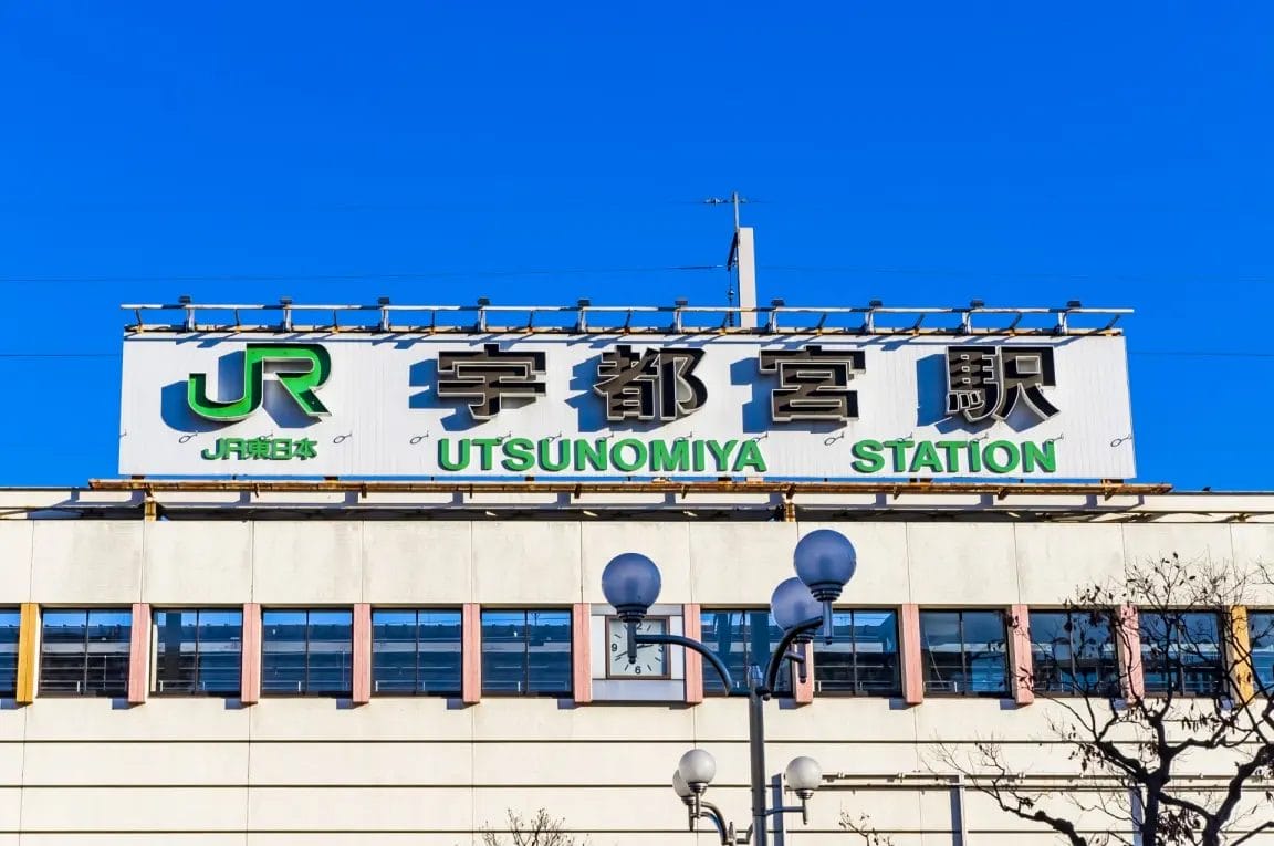 澄み切った青空の下、白いビルの上に日本語と英語の文字が書かれた宇都宮駅の看板。.