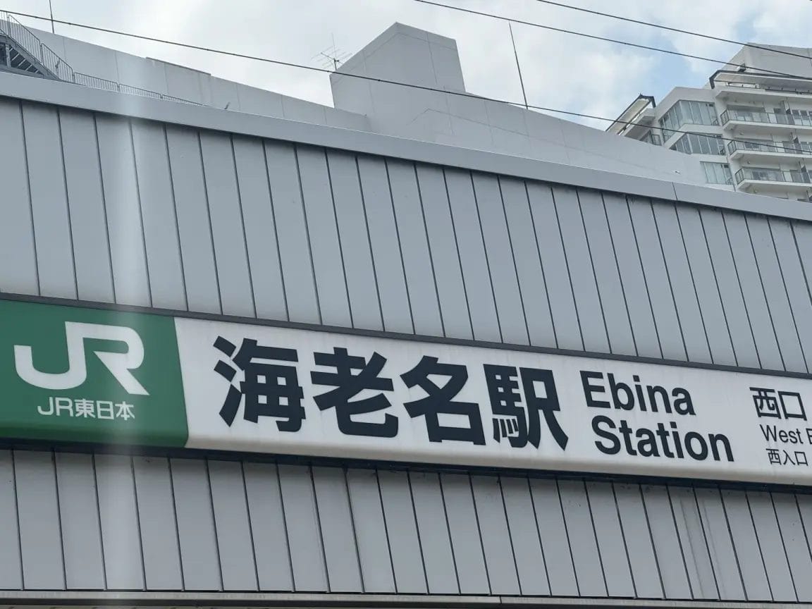 ビル入口上部の看板には、英語と日本語で「海老名駅」と表示され、JR東日本のロゴと西口への道順が記されている。.