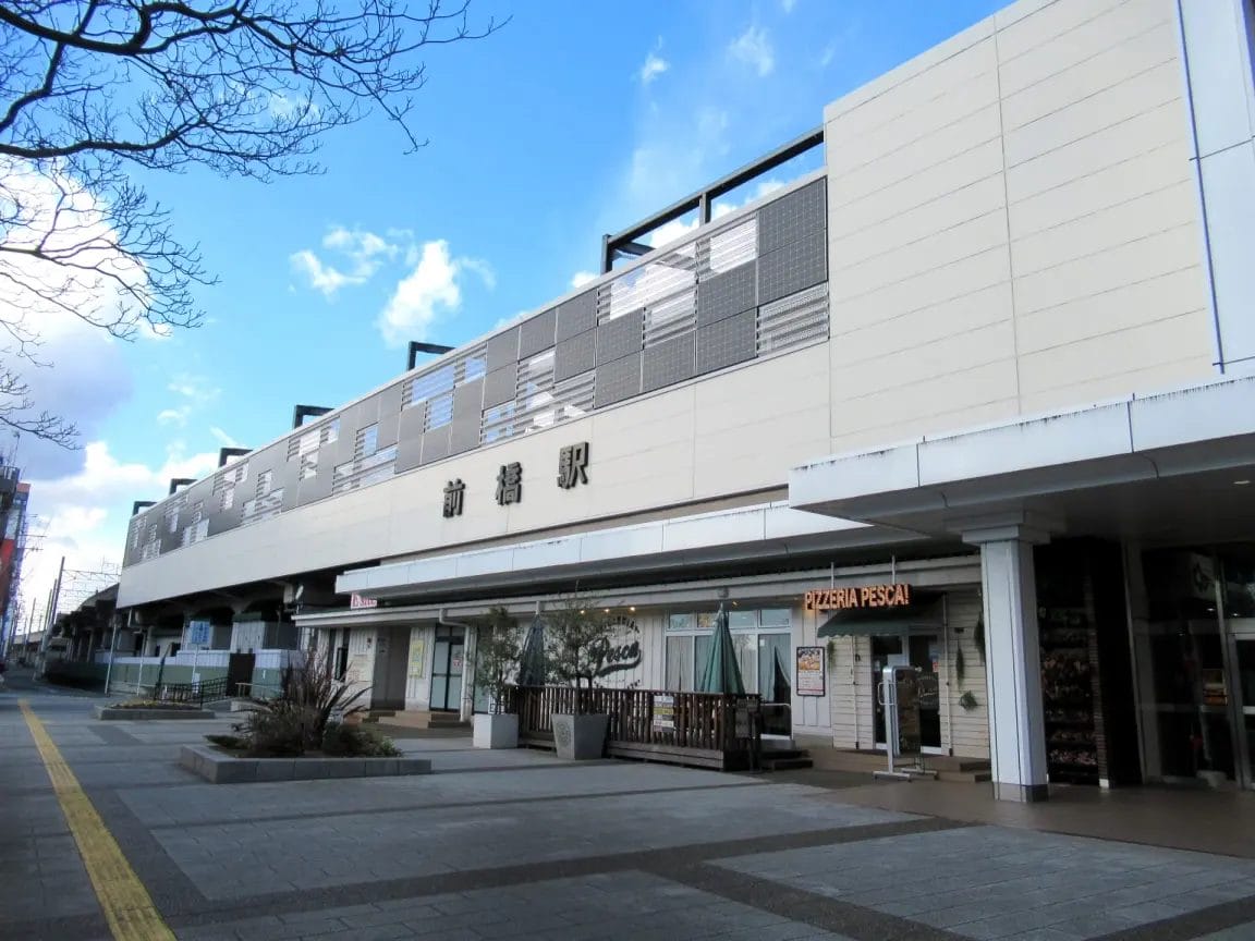 一部曇り空の下、ファサードには日本語の看板が掲げられ、路面には商店が並ぶモダンな明るい色の駅舎。.