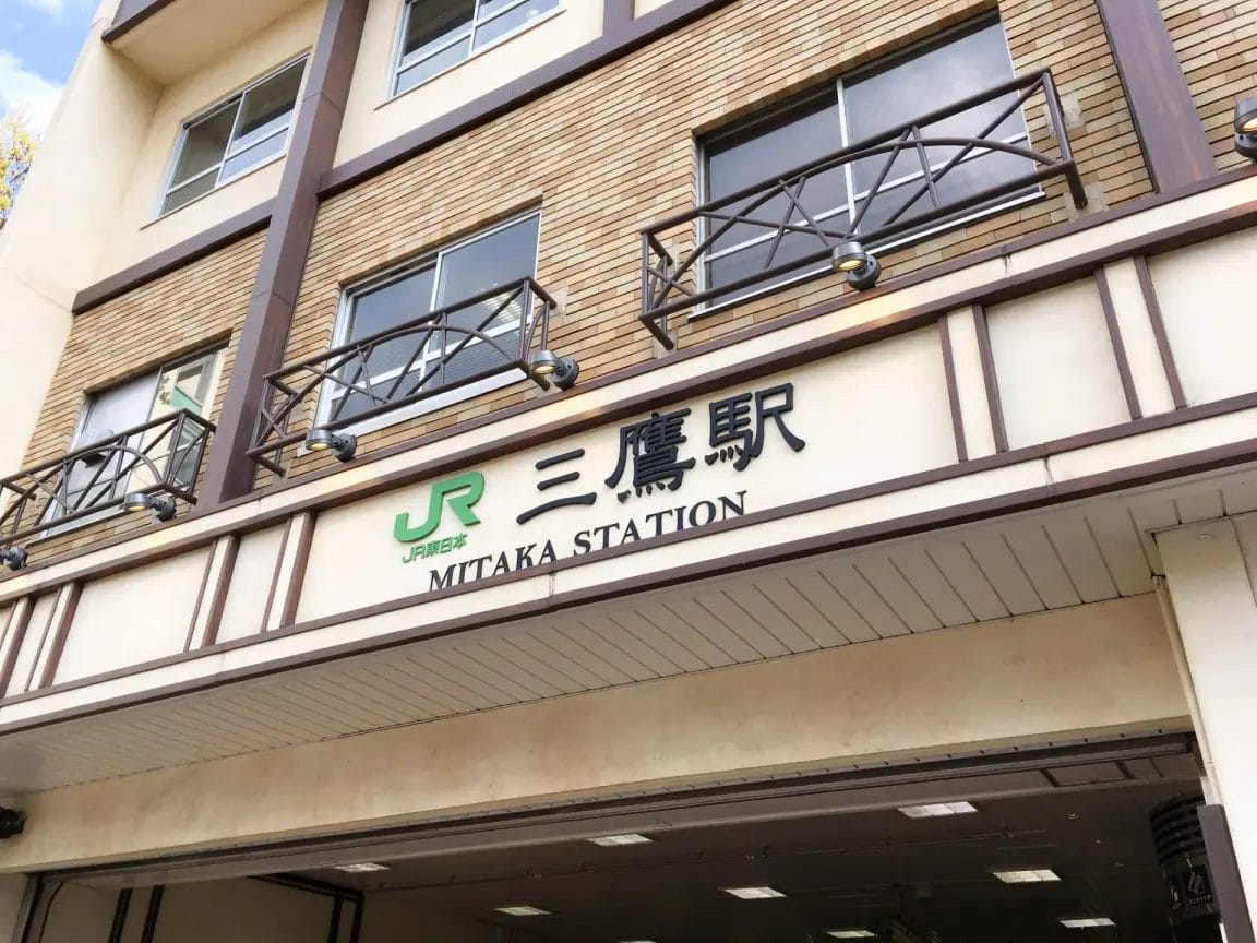 日本の三鷹駅のエントランス。JRのロゴと、ビルのファサードにある日本語と英語の看板が見える。.