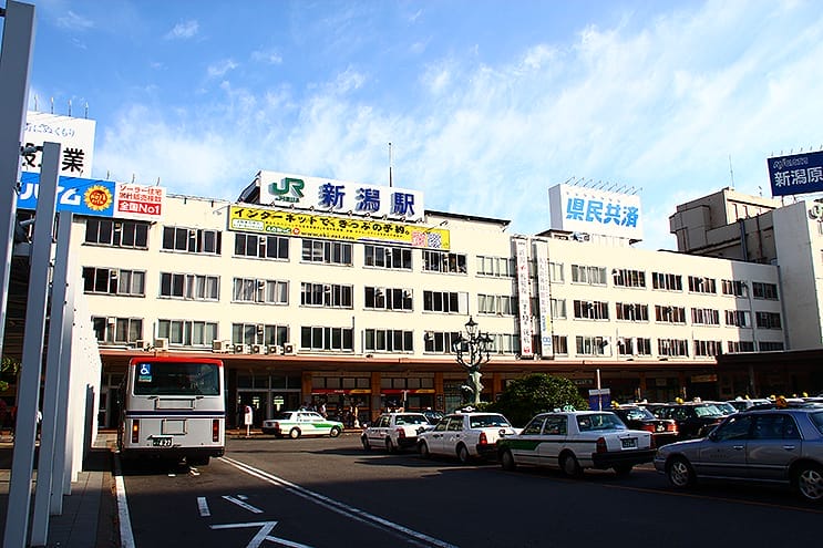 青空の下、新潟駅などの看板が掲げられた雑居ビルの前にバスやタクシー、車が停まっている。.