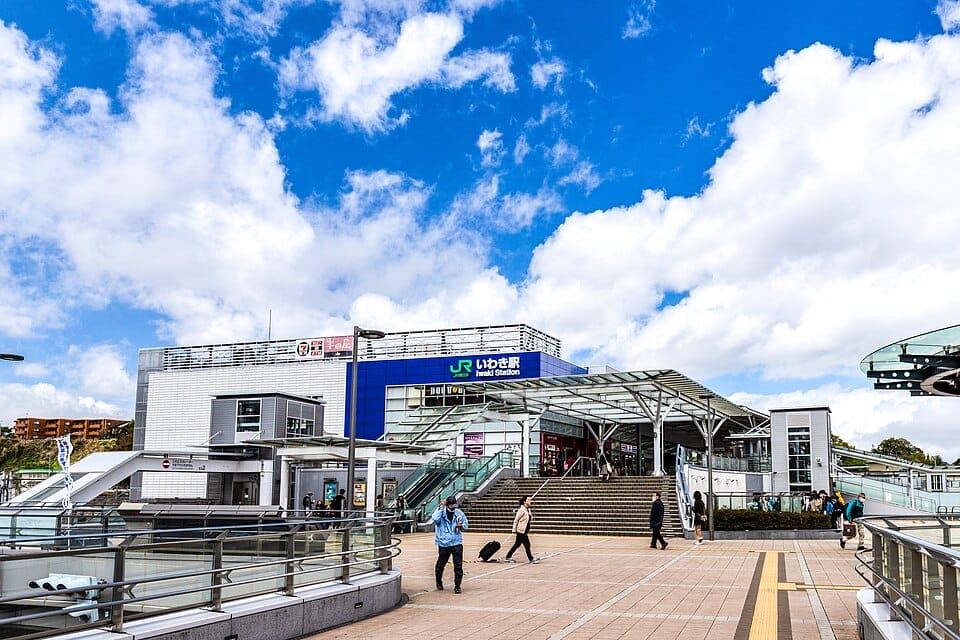 青いJRの看板、ガラス張りのエントランスのキャノピー、そして青空と散在する雲に覆われた晴れた日に歩く数人の人々。.