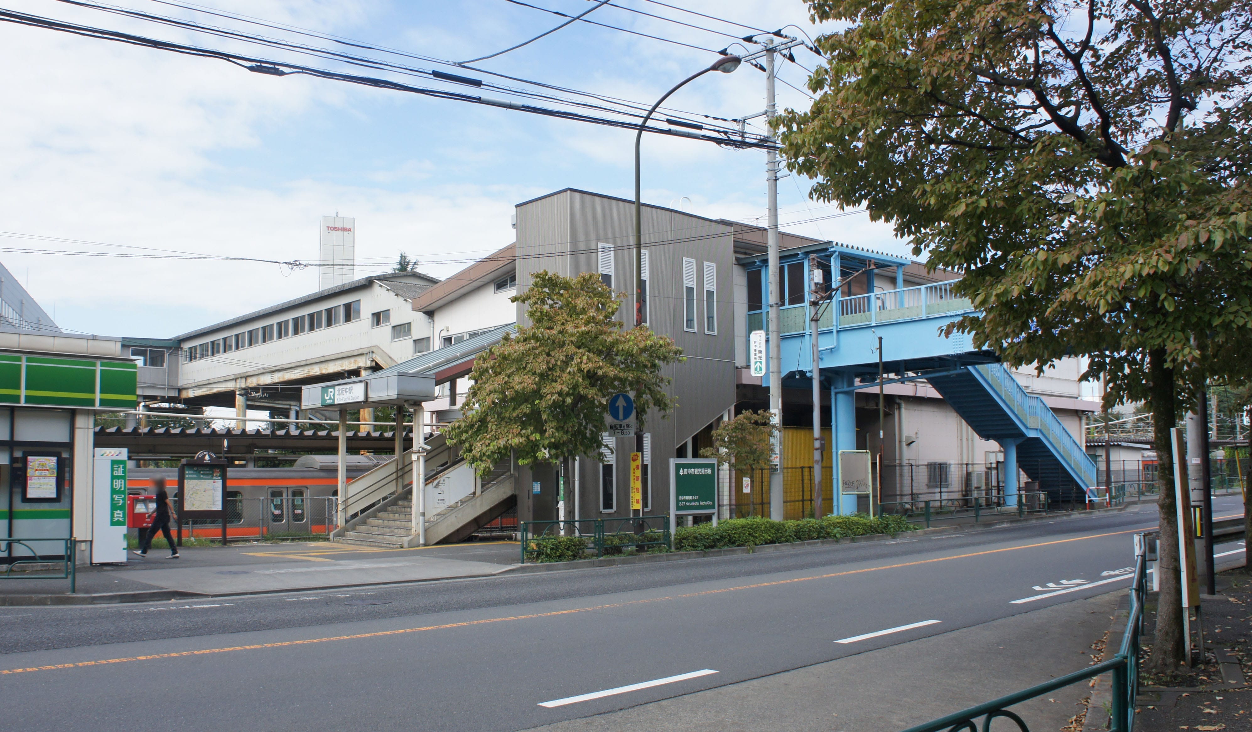 府中市にある北府中駅のストリートビュー。駅舎、線路、青い高架歩道が見える。.