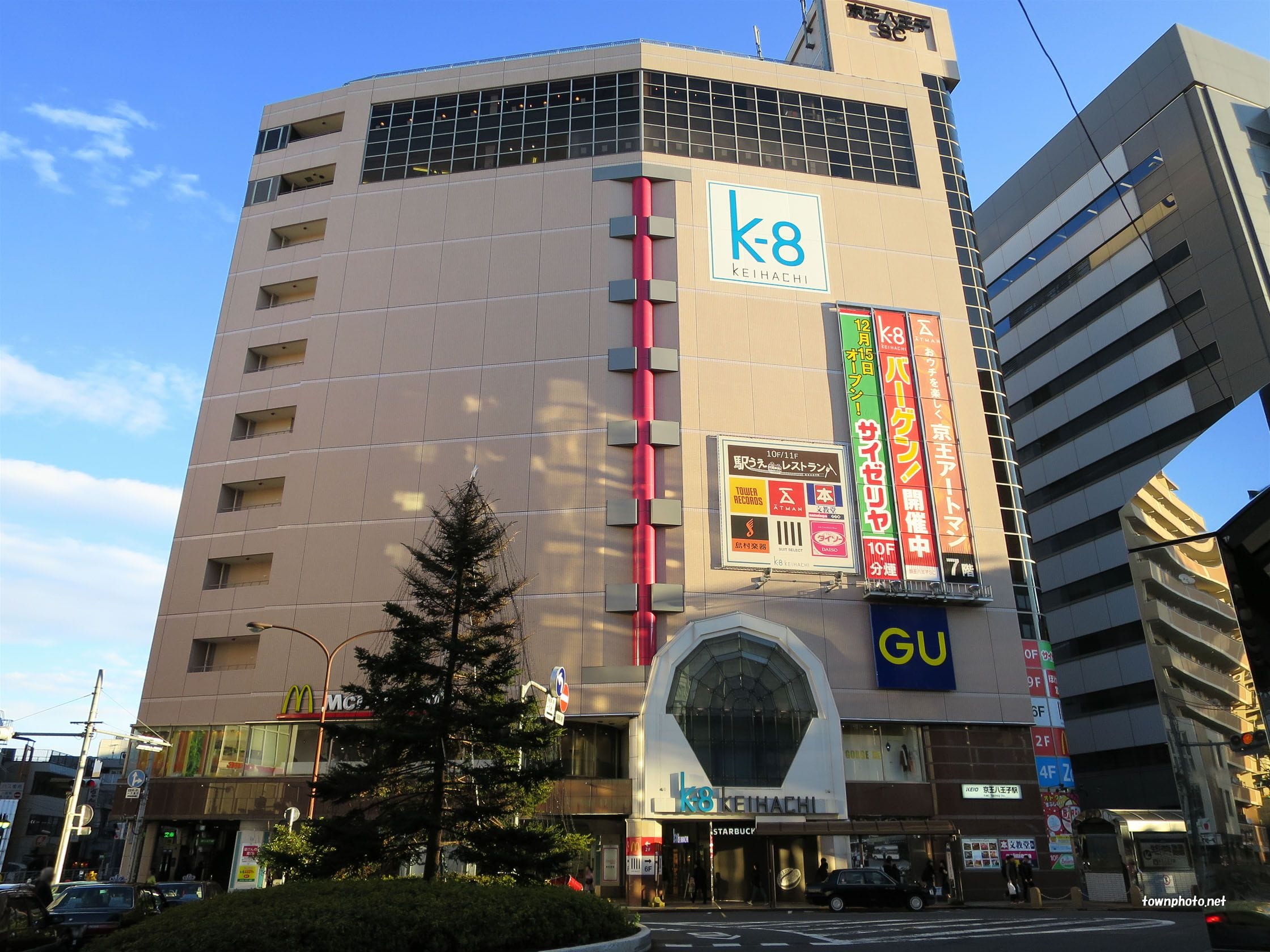都会の晴れた空の下、マクドナルドやGUなど様々な店舗の看板が並ぶ立体的な商業ビル。.
