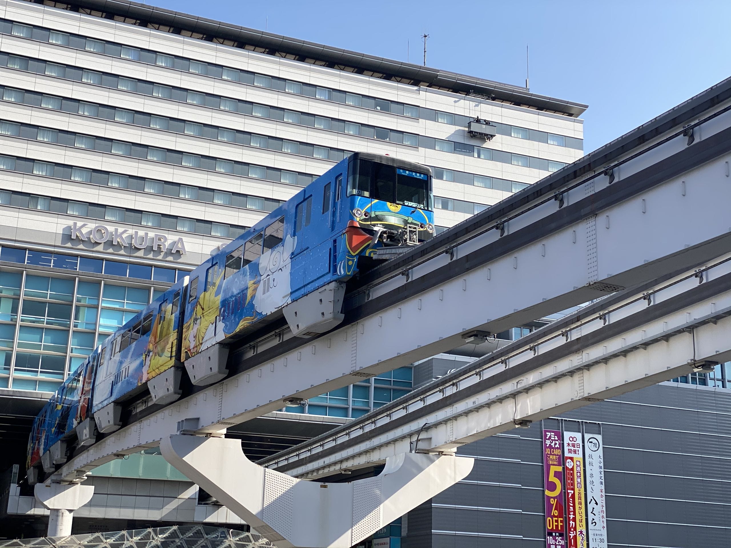 都会的な晴天の下、カラフルなアートワークが施された青いモノレールが小倉駅舎前を走る。.