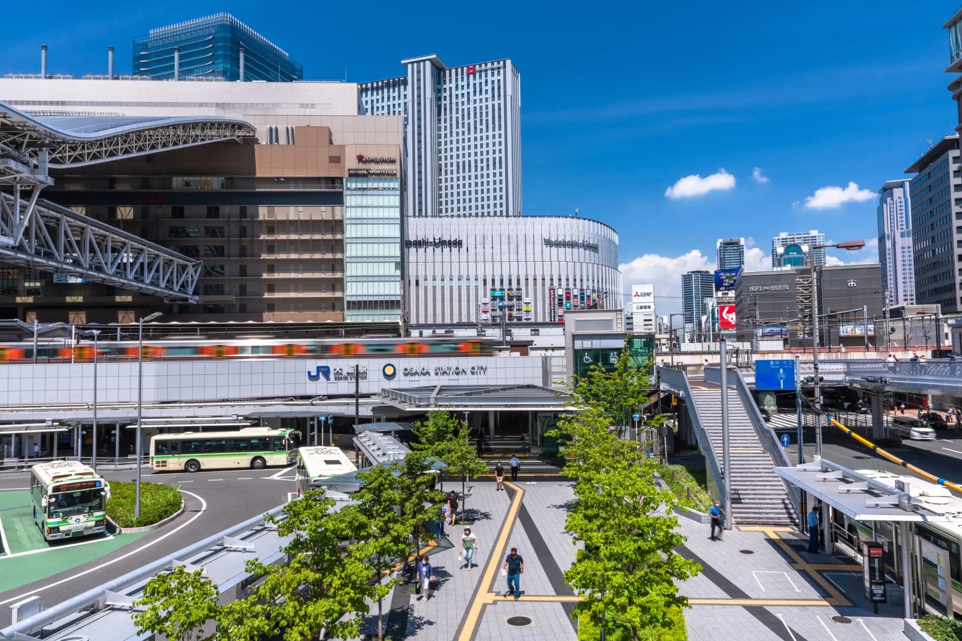 澄み切った青空の下、行き交う電車、バス、歩く人々、高層ビル。.