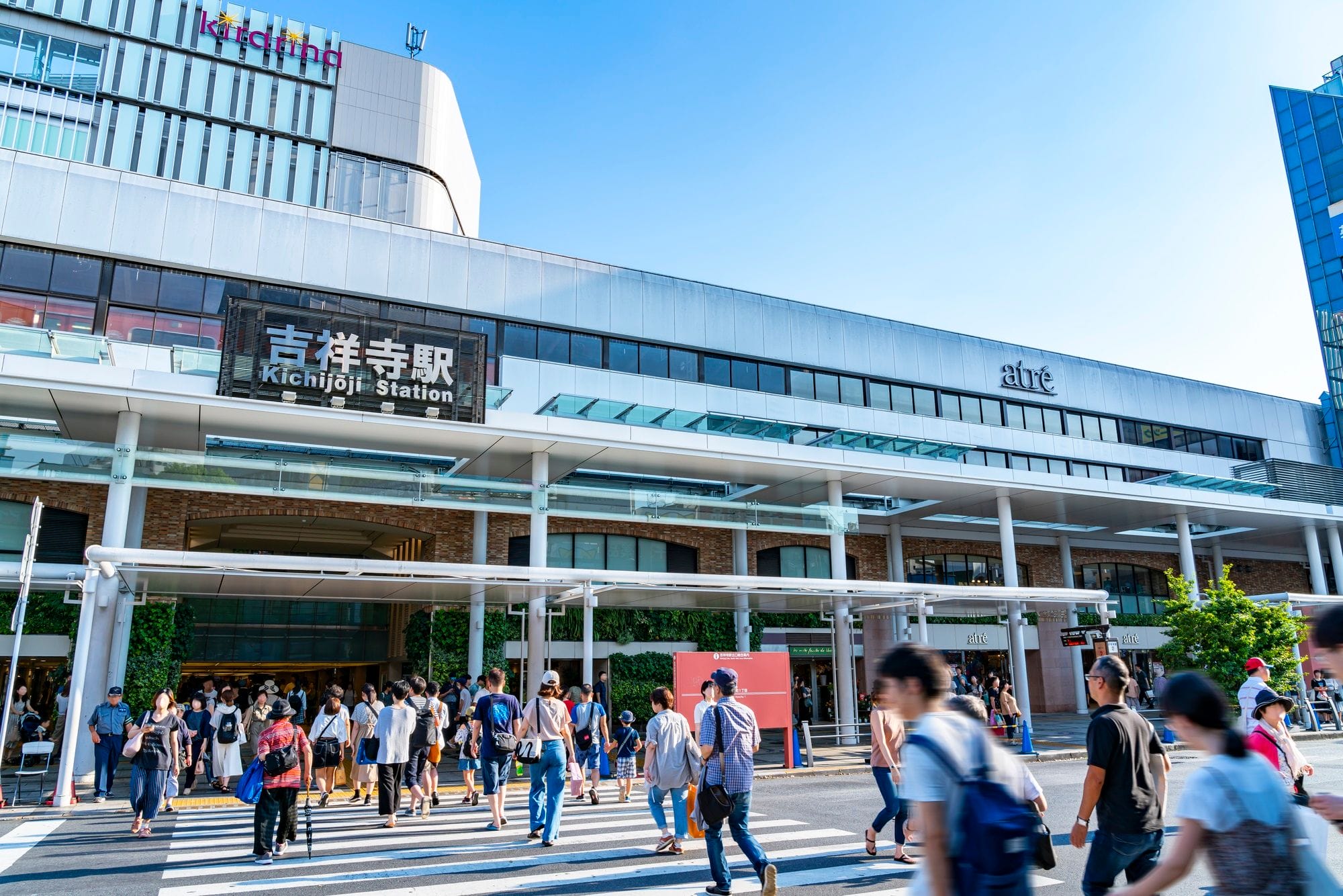 晴れた日の東京・吉祥寺駅前を歩く人々の群れ。駅ビルには店舗や看板が見える。.