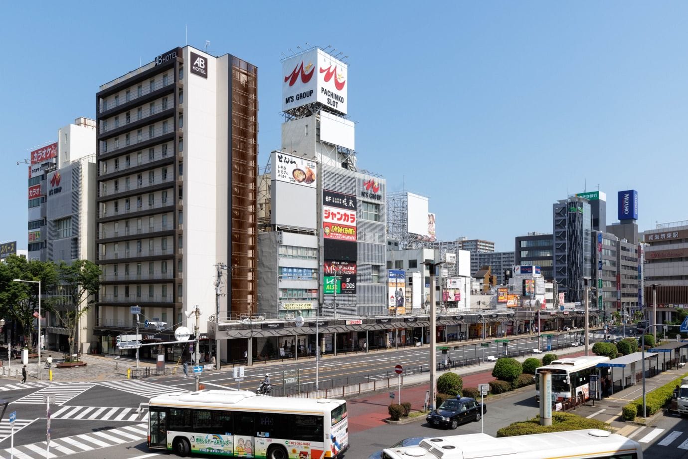 様々な標識が表示された高層ビル、手前に停車中のバス、そして数人の歩行者と車が行き交うほとんど人通りのない道路。.