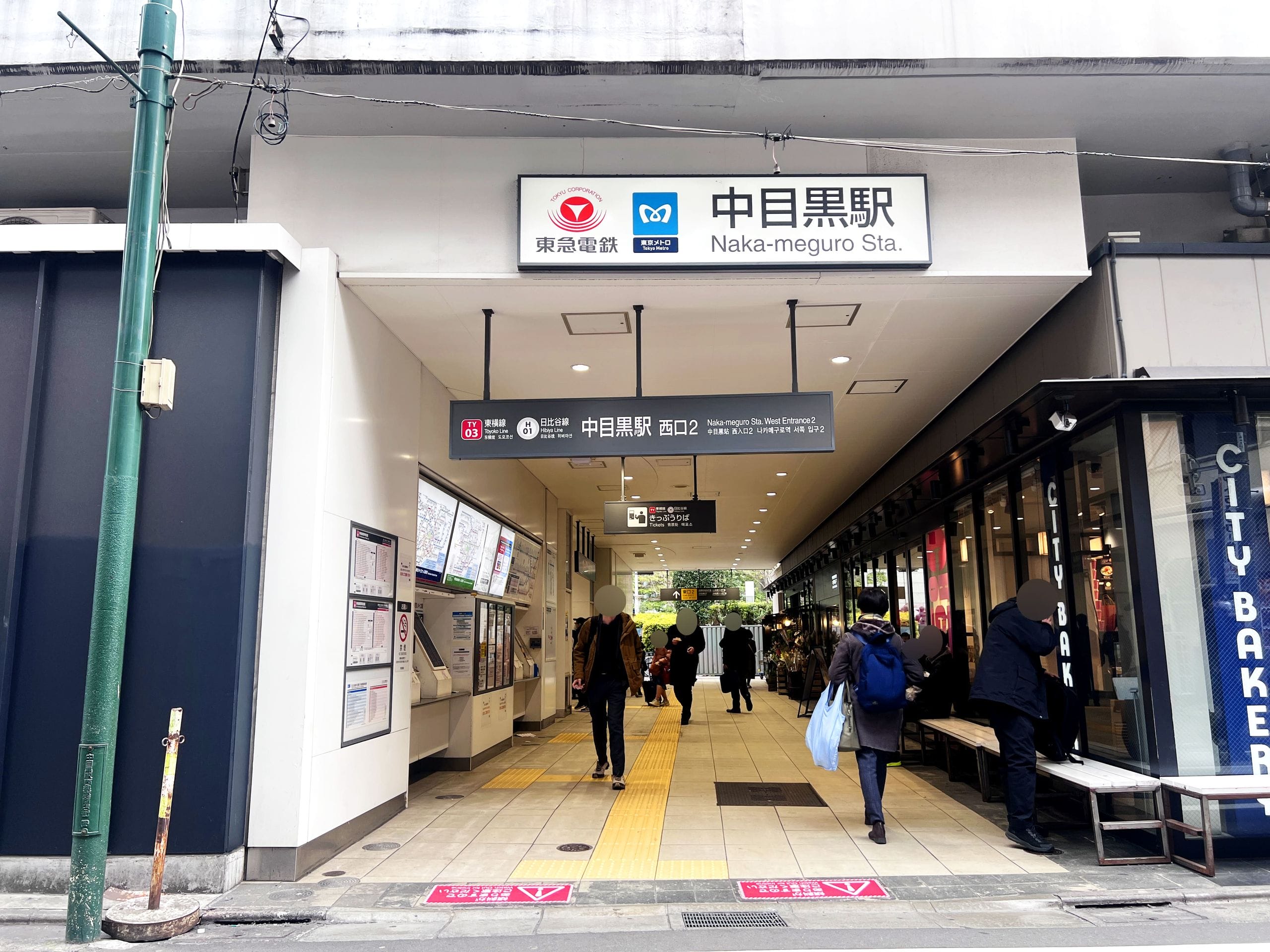 東京・中目黒駅の入り口。出入りする人々、上の看板、券売機、右側のパン屋。.