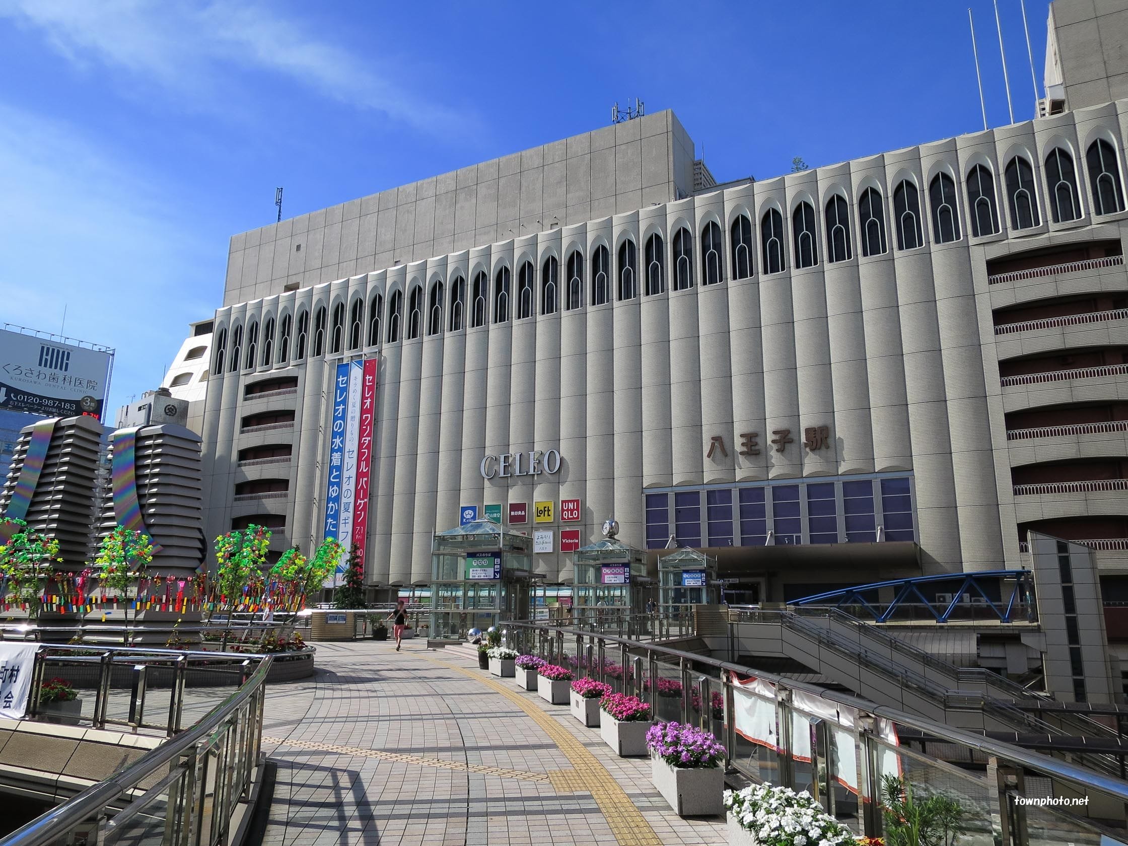 八王子駅のモダンなコンクリート・ファサードのワイド・ビュー。西武の看板、花のプランター、そして上空には澄み切った青空が広がる。.