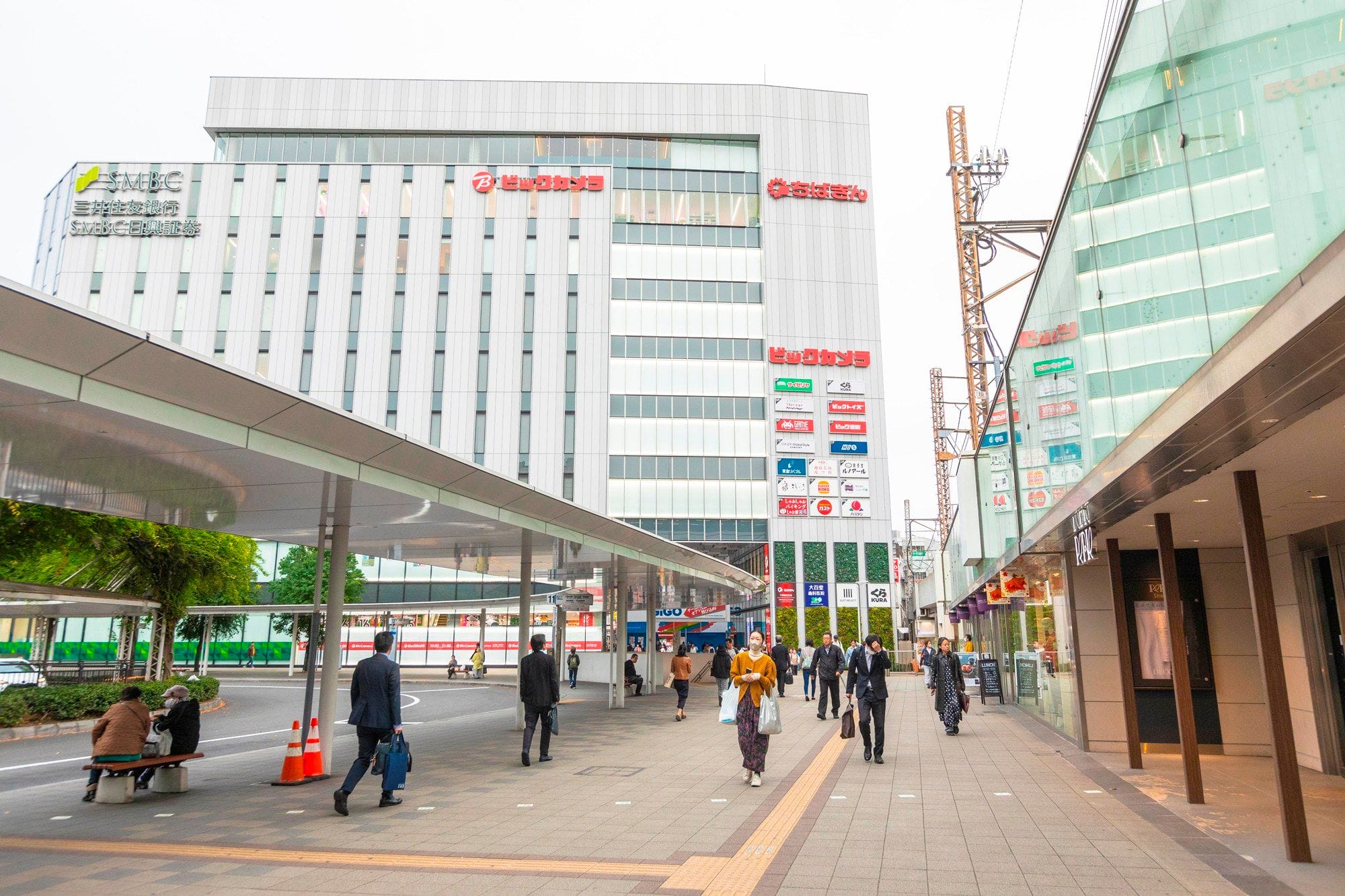 都市部の曇り空の下、日本語の看板がいくつも掲げられた近代的なビルの横を、広い歩道を歩く人々。.