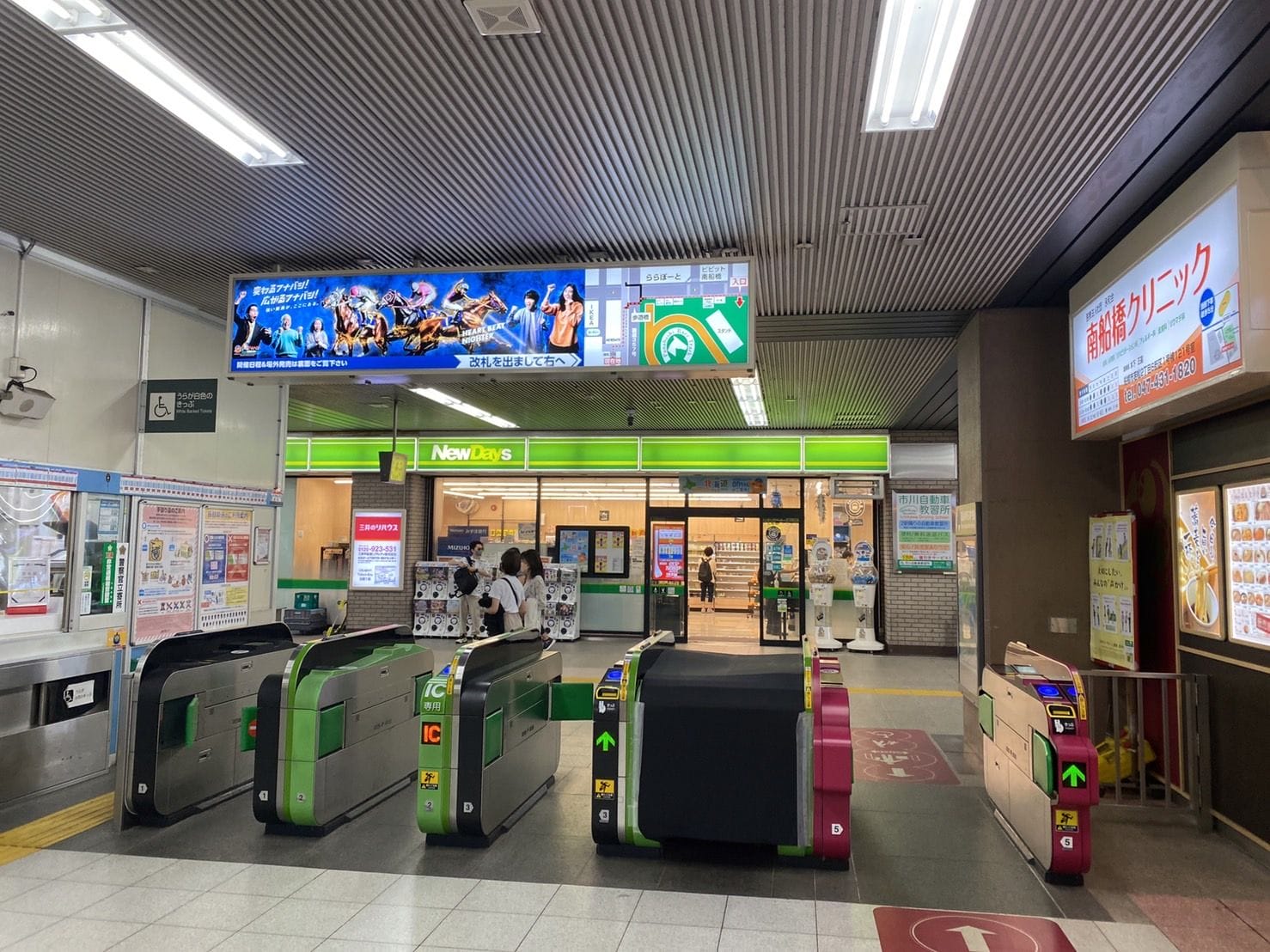 日本の駅の改札口。コンビニエンスストアと中にいる人々、照明付きの看板、頭上の広告が見える。.