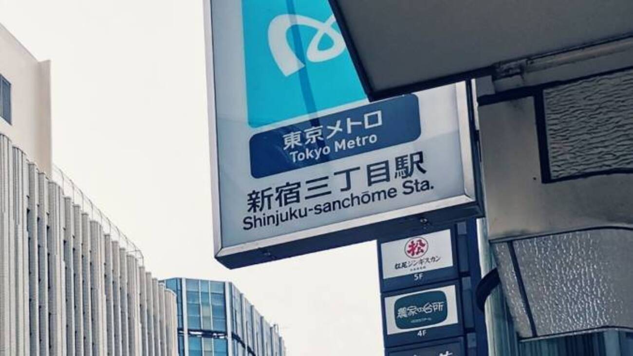 東京メトロ新宿三丁目駅の街路標識。背景には近隣のビル看板と近代建築。.